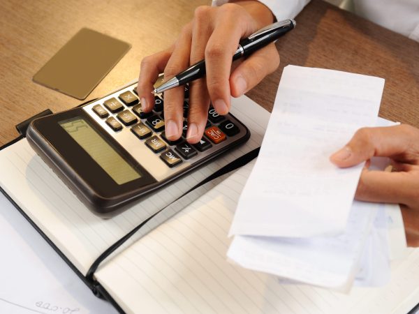 Closeup of person holding bills and calculating them. Notebook, calculator and credit card lying on desk. Payment concept. Cropped view.