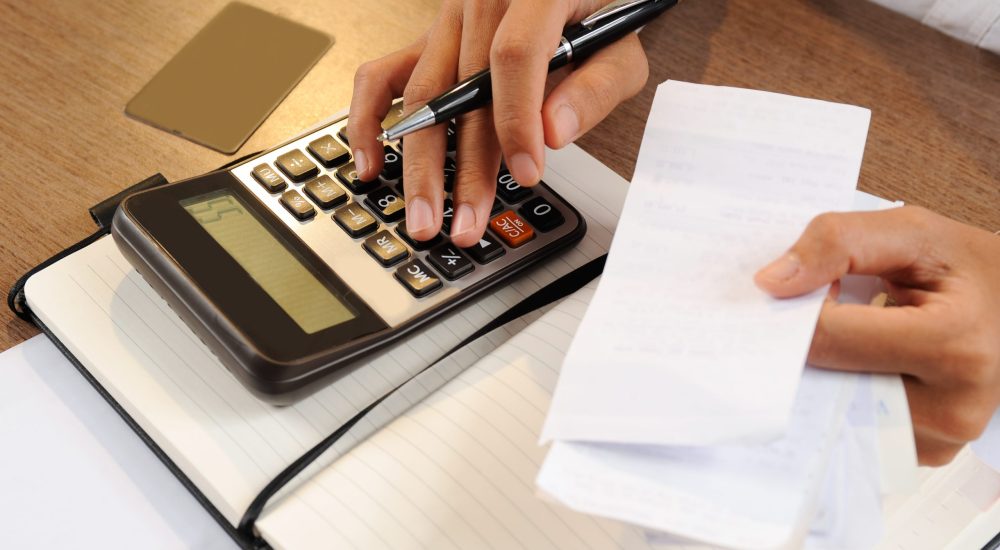 Closeup of person holding bills and calculating them. Notebook, calculator and credit card lying on desk. Payment concept. Cropped view.