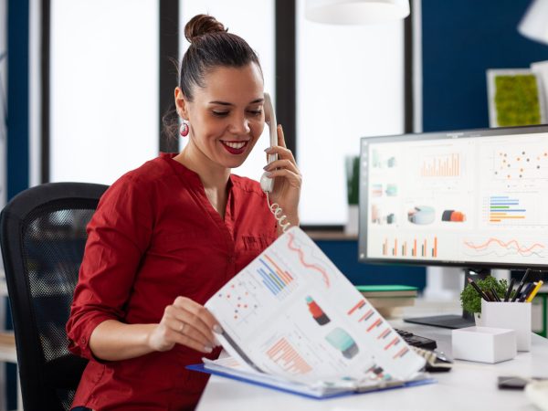 Businesswoman in company office checking finaicial statistics on clipboard, discusing details with coworkers during phone call conversation. Happy entrepreneur communicating with client.