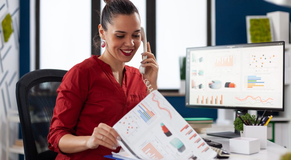 Businesswoman in company office checking finaicial statistics on clipboard, discusing details with coworkers during phone call conversation. Happy entrepreneur communicating with client.