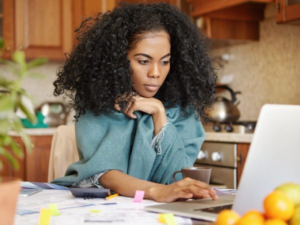 Beautiful young dark-skinned woman with Afro hairstyle wearing wrap keeping hand on touchpad of open laptop computer , looking at screen with serious concentrated expression, paying bills online