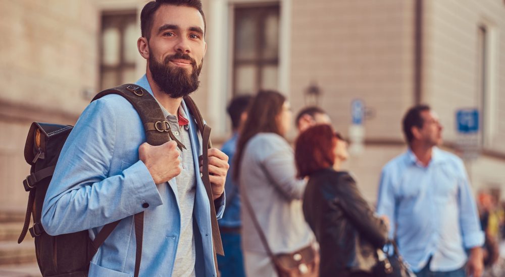 A tourist with a full beard and haircut, wearing casual clothes, holds a backpack and texting on a smartphone, standing on an antique street, during the excursion in Europe.