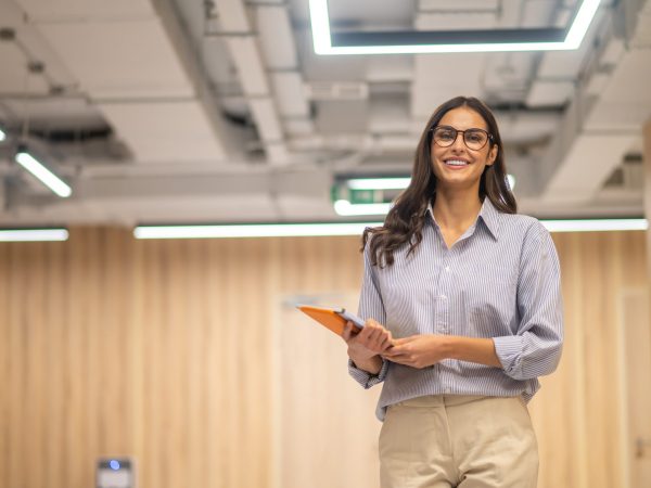 Good mood. Young long haired woman in glasses holding folder smiling at camera in illuminated room