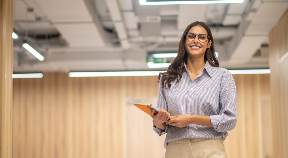 Good mood. Young long haired woman in glasses holding folder smiling at camera in illuminated room